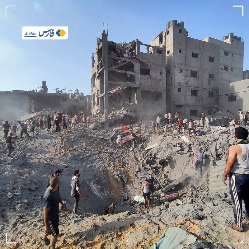 People gather among the rubble and dust around a heavily damaged multi-story concrete building, with a large crater visible in the foreground.