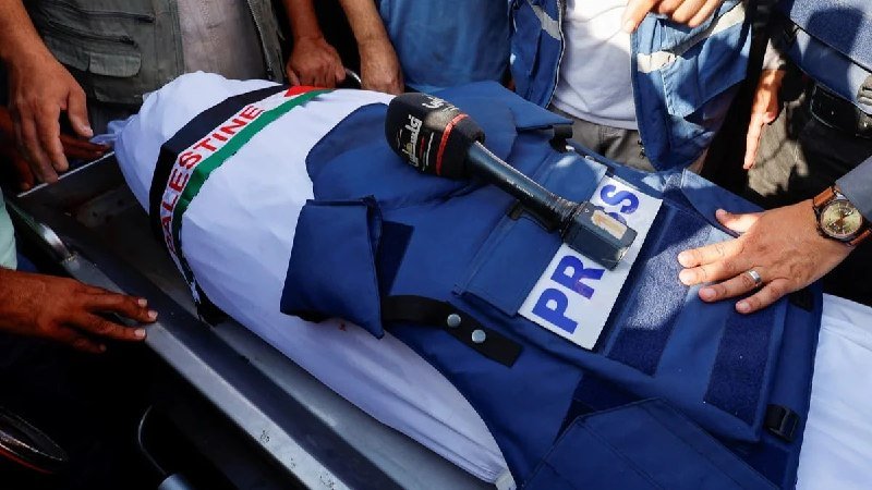 A body draped in a Palestinian flag lies on a stretcher as uniformed personnel and others in civilian clothing stand around, one wearing a "PRESS" vest with a microphone resting on it.