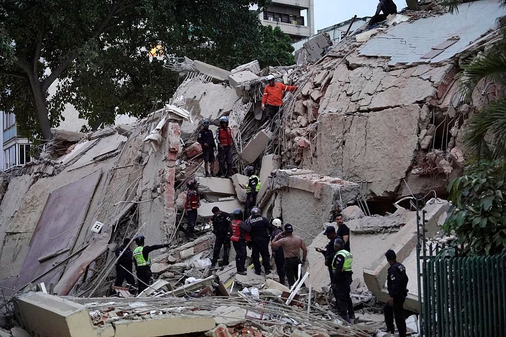 Rescue workers in helmets and high-visibility vests climb through the rubble of a collapsed concrete building, searching the debris.