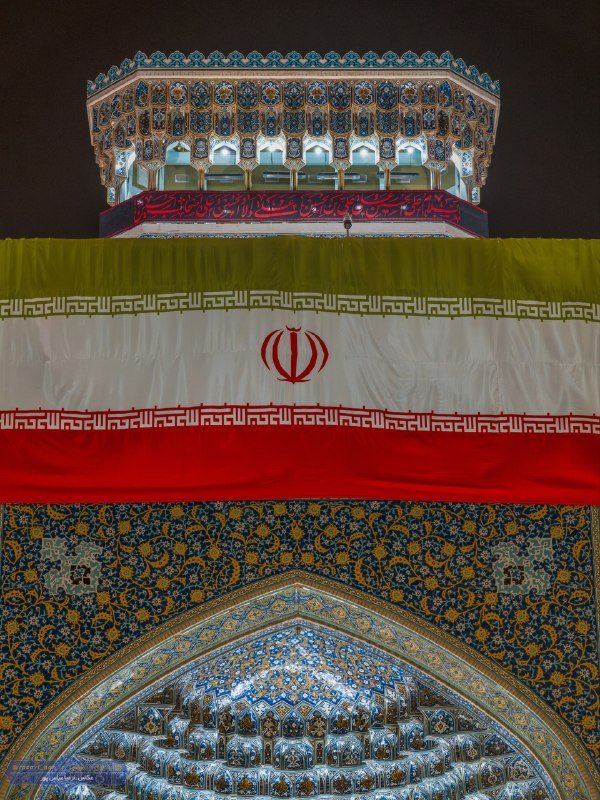 The illuminated tilework of an ornate Islamic shrine dome is draped with a large Iranian flag featuring its emblem, photographed at night.