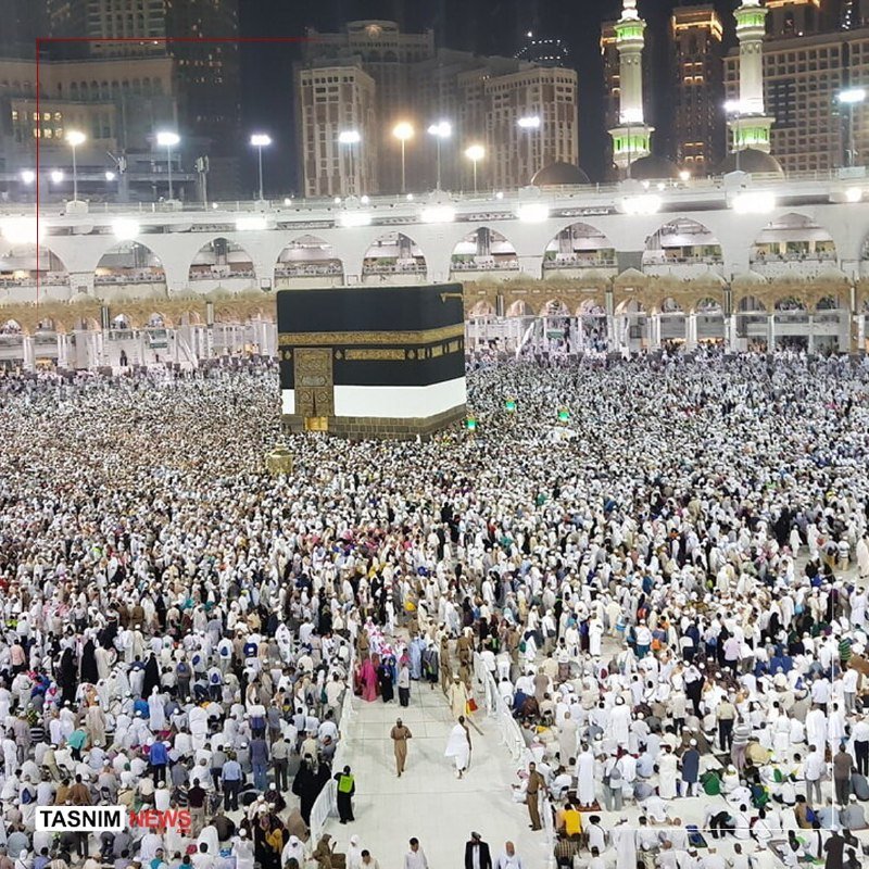 A vast crowd in white garments circles a large black cubic structure within a brightly lit mosque complex at night, with illuminated minarets and high-rise buildings visible in the background.