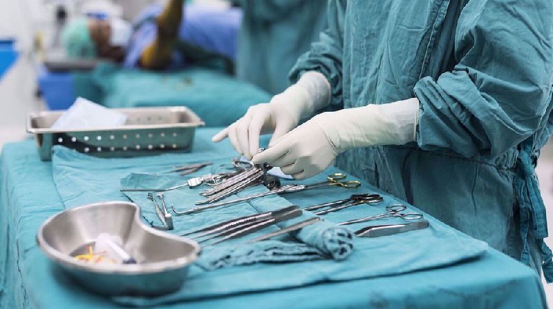 A medical professional wearing scrubs and white gloves handles surgical instruments, including scissors and clamps, laid out on a teal-draped table.