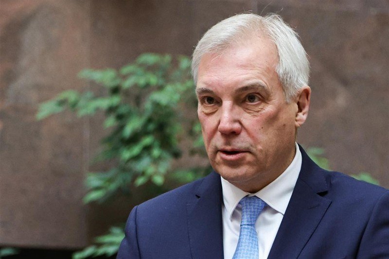 A gray-haired man in a dark suit and light blue tie speaks indoors, with green plants and a stone wall visible behind him.