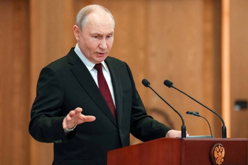 A gray-haired man in a dark suit and red tie speaks at a wooden podium with microphones, gesturing with his hand in a wood-paneled room.