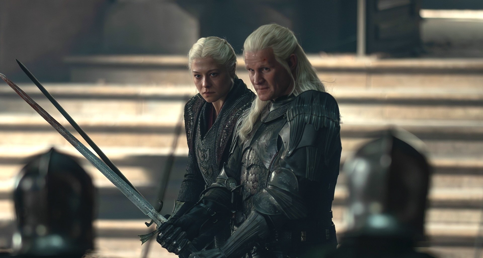 Two platinum-haired figures in dark medieval armor stand on stone steps, one holding a long sword, with armored horses visible behind them.