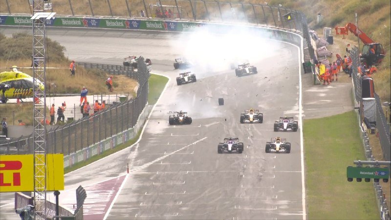 Multiple open-wheel race cars navigate a track following a collision, with smoke rising and debris scattered across the circuit as track marshals in orange suits respond along the barriers.