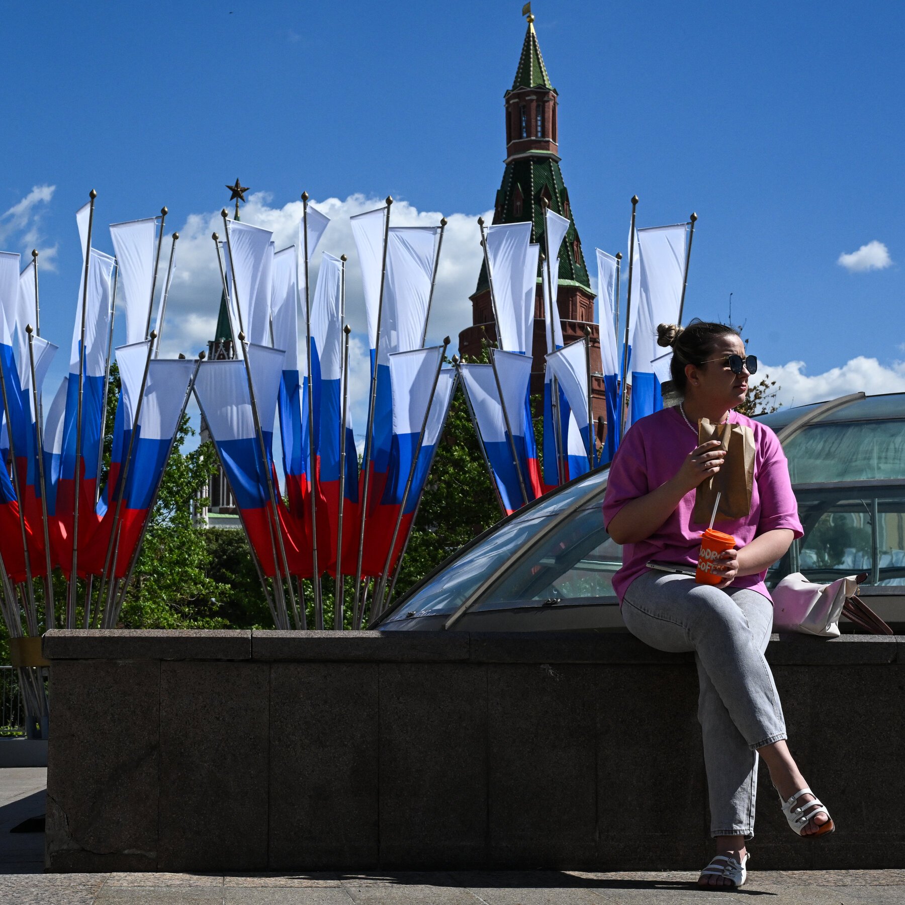 A woman in a pink shirt and sunglasses sits on a stone ledge holding a paper bag and orange cup, with Russian flags and a red-brick Kremlin tower in the background.