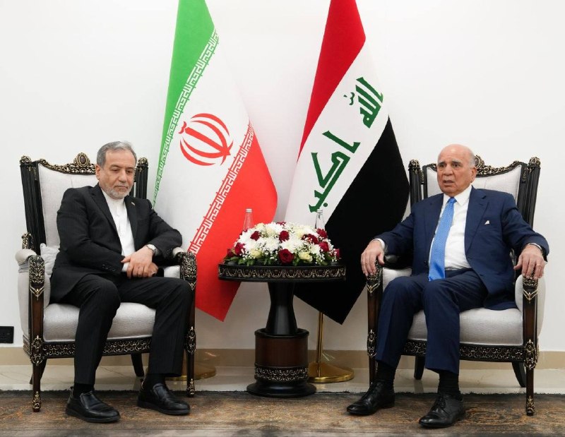 Two men sit facing each other in an ornate wood-paneled room with an Iranian flag and bookshelves, surrounded by seated groups of men taking notes.