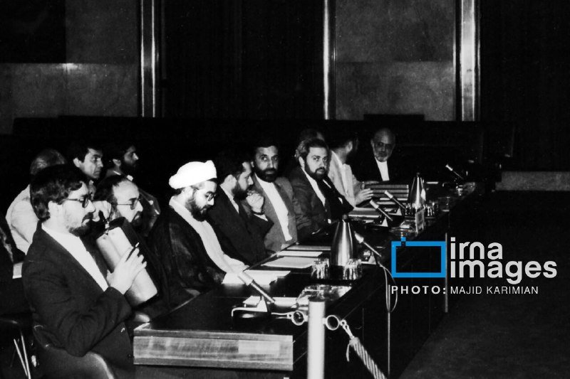 Black-and-white photo shows several men in suits seated at a long conference table with microphones, including a cleric in a white turban, appearing to participate in a formal meeting.