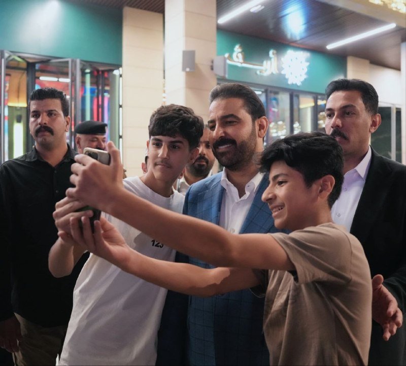 A bearded man in a blue suit jacket takes a selfie with a smartphone alongside smiling youths and suited men outside a storefront with Arabic signage at night.