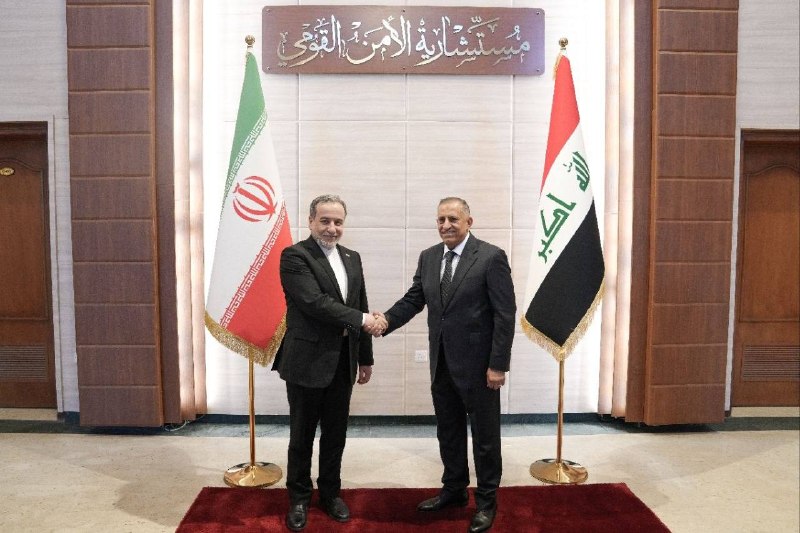 A group of men seated on couches face two men seated centrally in an ornate, wood-paneled room featuring a chandelier, bookshelves, and an Iranian flag.