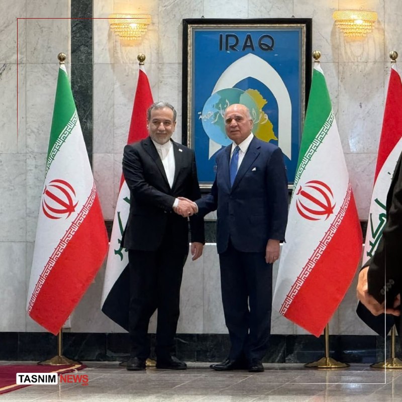 A group of men in dark clothing sit in a wood-paneled meeting room facing each other, with an Iranian flag and two framed portraits displayed on the back wall.