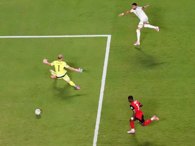 A soccer ball rolls past a diving goalkeeper in yellow toward the goal line as a player in red watches and a defender in white sprints nearby.