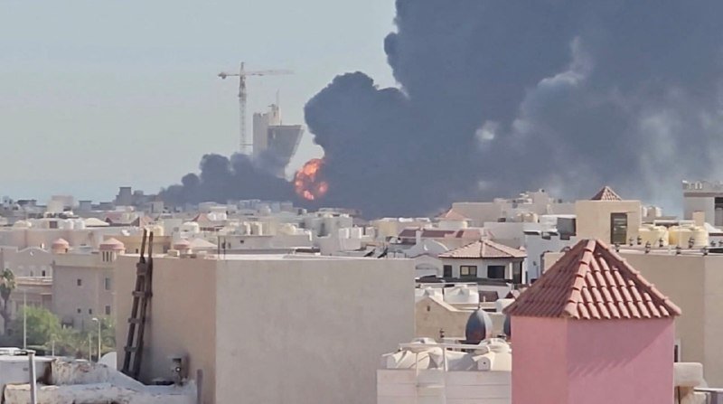 Large black smoke plume and visible flames rise over a city of densely packed light-colored buildings, with a construction crane visible in the background.