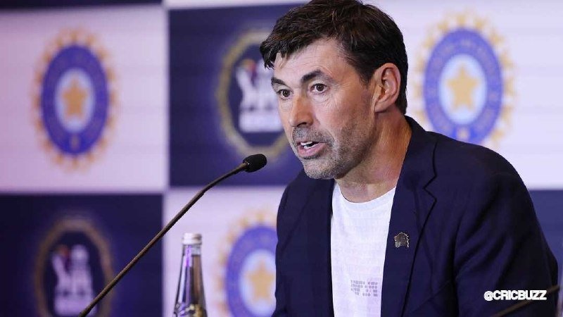 A man in a dark blazer speaks into a microphone during a press conference, with a backdrop featuring circular logos.