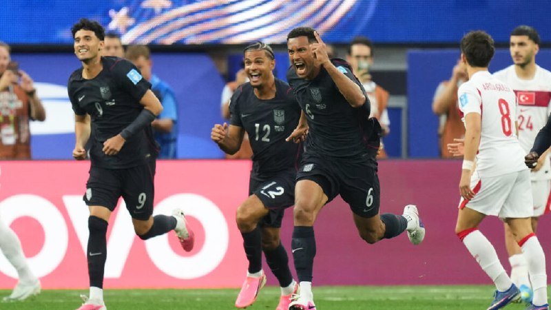 Three soccer players in black England jerseys celebrate exuberantly on a pitch, while a player in a white and red Turkey kit stands nearby.