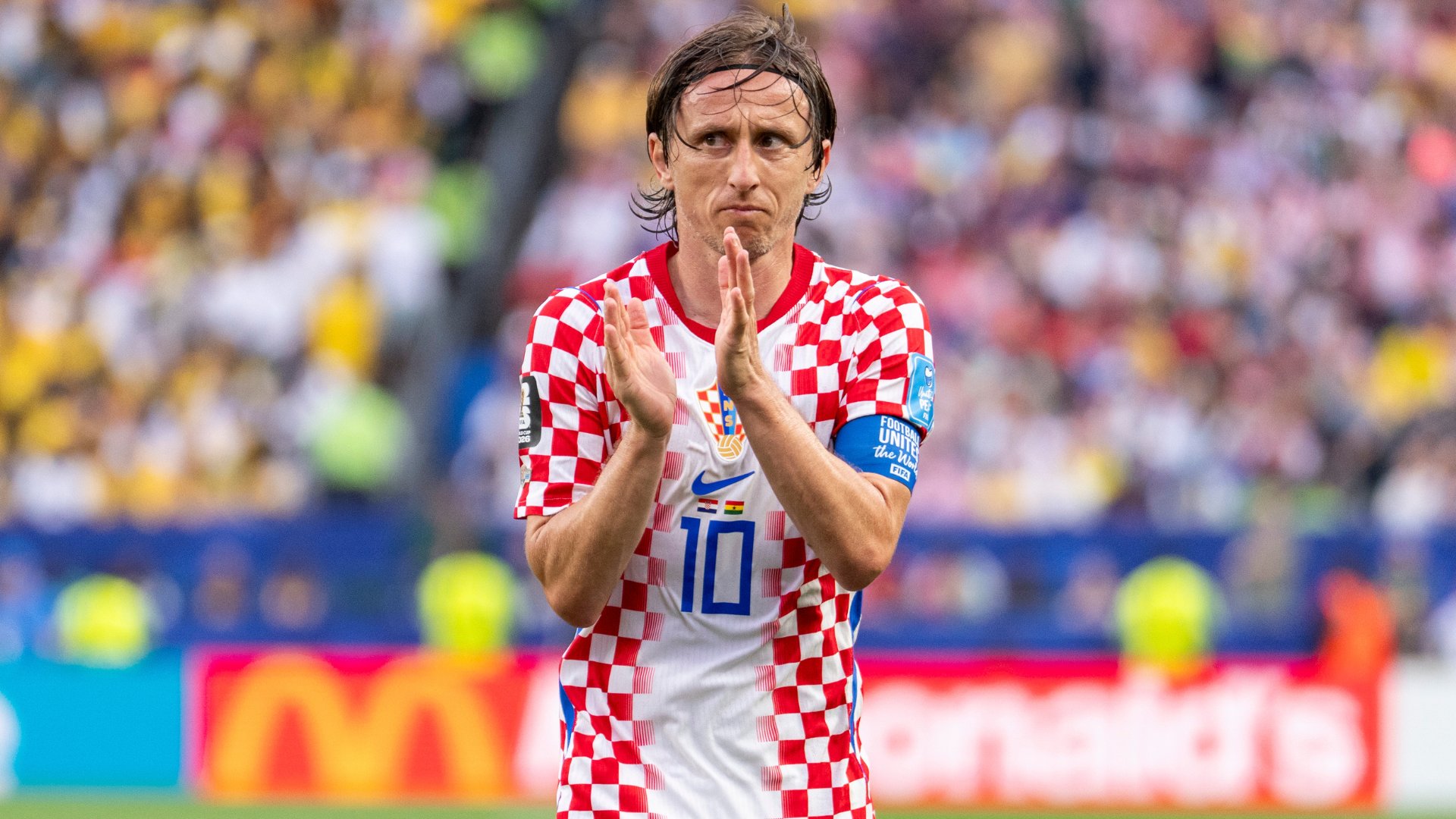 A soccer player wearing a red and white checkered jersey with the number 10 applauds in a stadium filled with spectators.