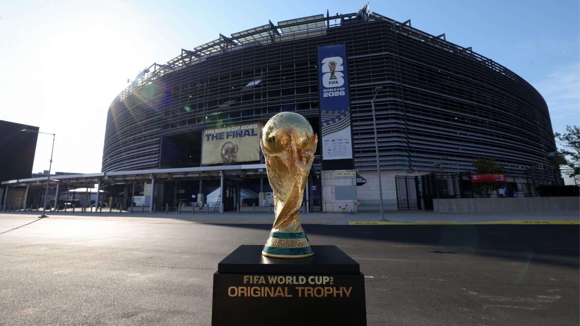 A golden FIFA World Cup Original Trophy replica sits on a black pedestal outside a large stadium displaying "FIFA WORLD CUP 2026" banners.