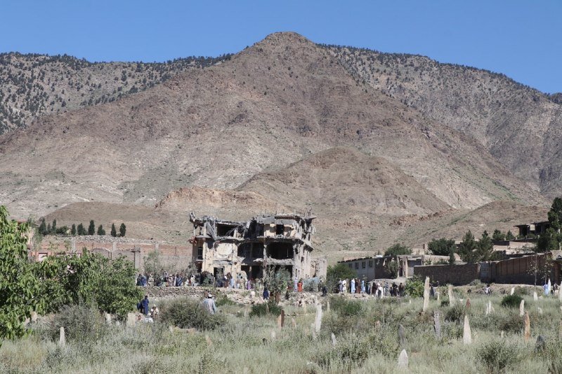 People gather near a heavily damaged multi-story concrete building, with small white flags marking graves in the surrounding field, set against a barren mountain under a clear blue sky.