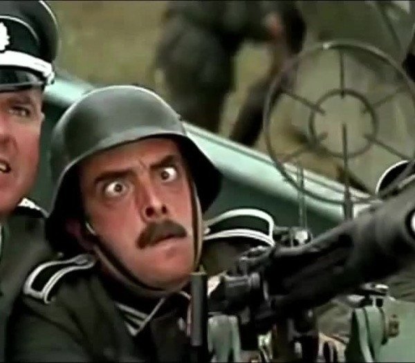 A wide-eyed soldier in a green helmet operates a large mounted machine gun, with a uniformed officer partially visible behind him.