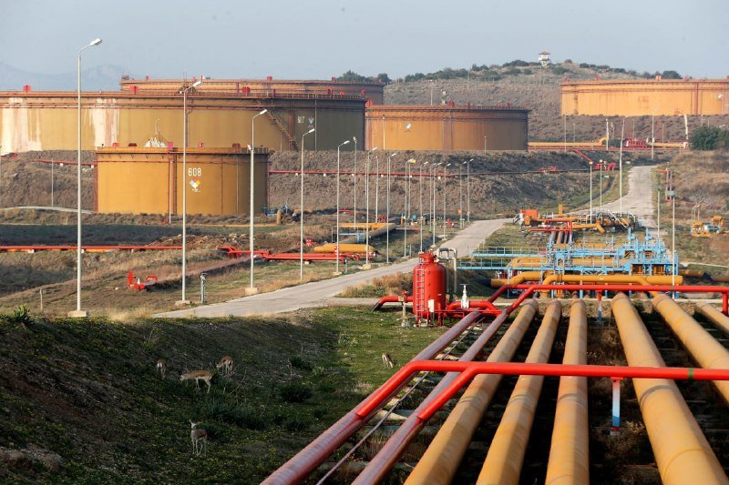 Large yellow industrial storage tanks and red and yellow pipelines stretch across a hillside, with a few deer grazing on the grassy foreground slope.