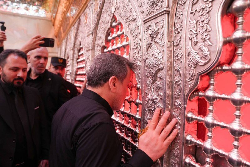 A man in a black shirt presses his face and hands against an ornate silver religious grille, while others behind him take photos with their phones.