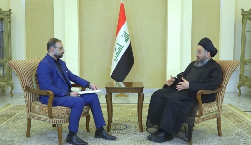 Two men sit in armchairs facing each other in a formal room, with an Iraqi flag positioned between them on a small table.