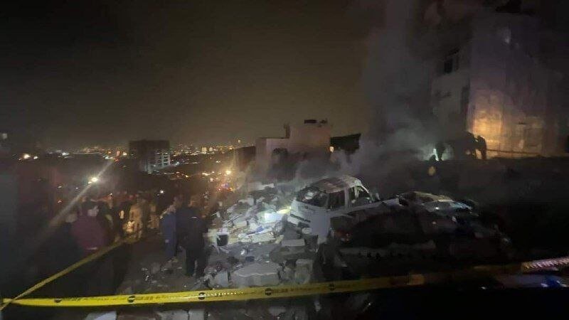 Numerous people, including responders, gather among concrete rubble beside a damaged vehicle behind yellow caution tape at night near a lit building.