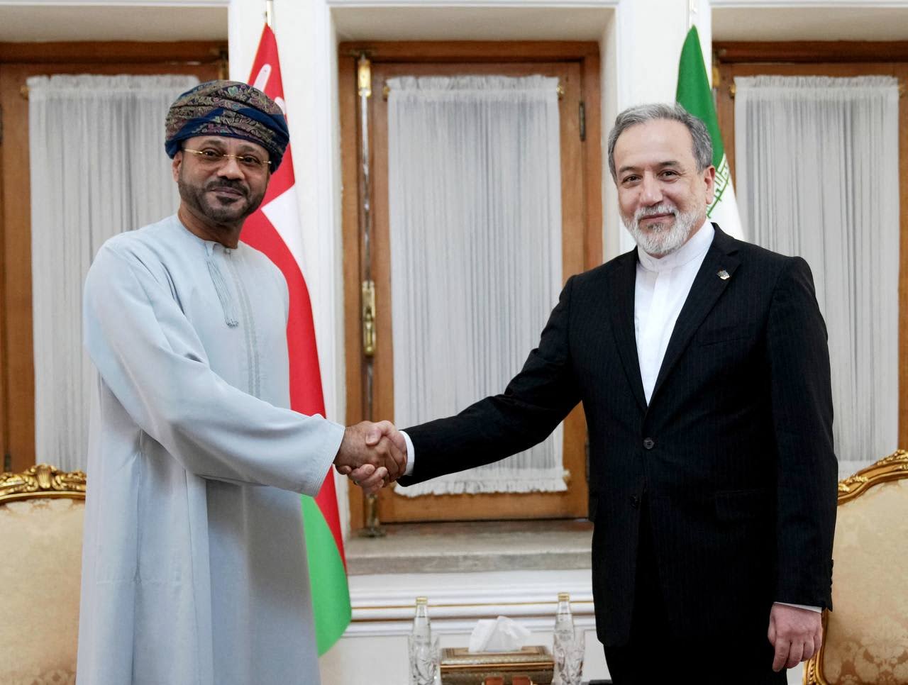 Two men shake hands indoors, one in a light blue traditional robe and patterned turban, the other in a dark suit, with Omani and Iranian flags visible behind them.