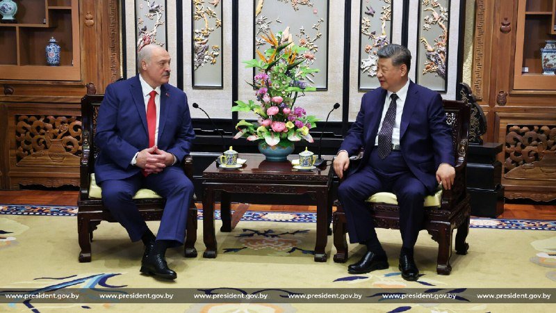 Two men in dark suits sit in ornate wooden chairs facing each other in a formal room with decorative panels, separated by a table holding a floral arrangement and teacups.