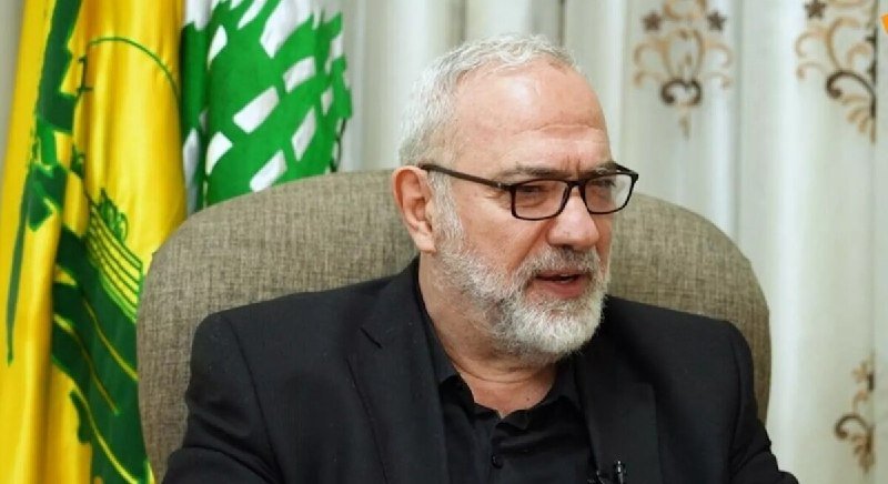 A bespectacled man in a dark suit speaks into a microphone at a podium, with a large banner featuring Arabic text and a portrait of another bearded man displayed behind him.