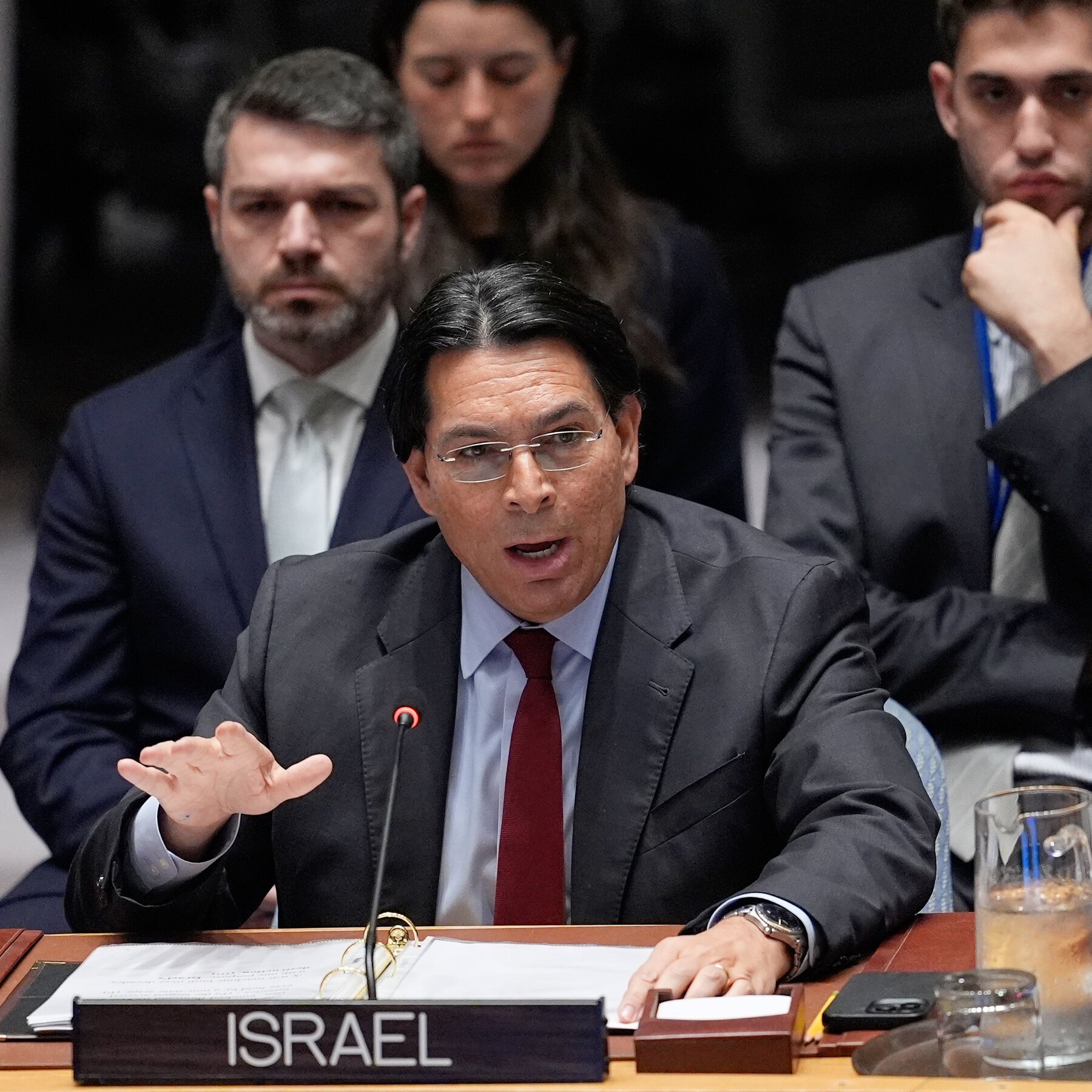 A man in a dark suit and red tie gestures while speaking into a microphone behind a placard reading "ISRAEL," with several other suited men seated behind him.
