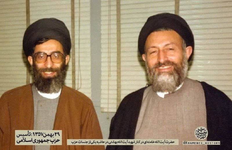 Two bearded men wearing black turbans and clerical robes stand smiling side by side in front of closed window blinds.