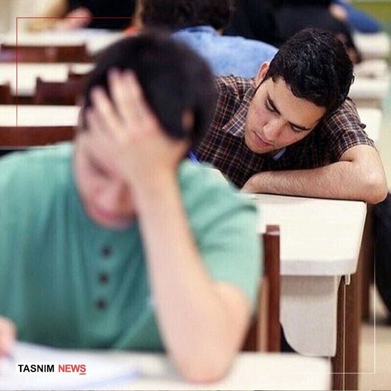 Young men sit at desks, one resting his head in his hand while another leans forward with his arms on the desk in what appears to be an exam setting.