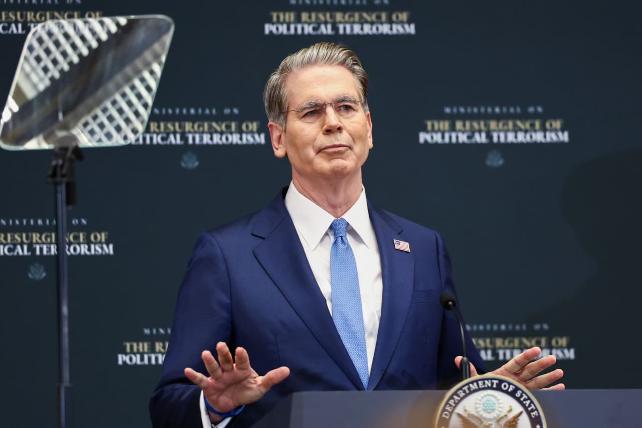 A gray-haired man in a navy suit and light blue tie stands at a podium gesturing with both hands at a State Department ministerial on political terrorism.