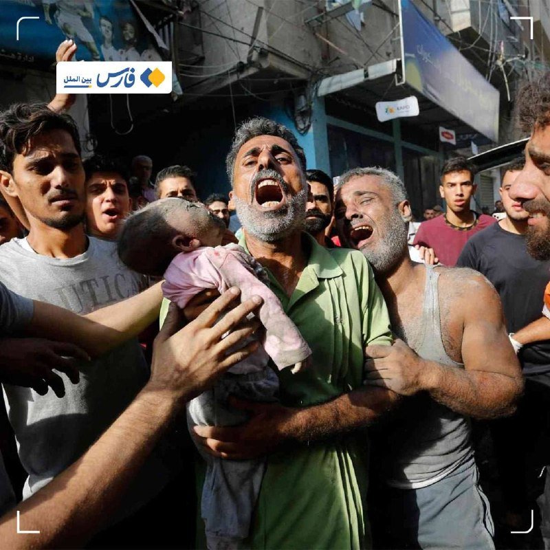 A group of men gathered on a street grieve while one holds a small child, with storefronts and Arabic signage visible in the background.