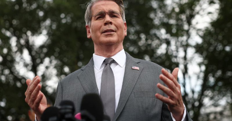 A man in a gray suit and tie with a small American flag pin stands outdoors gesturing with both hands raised, speaking in front of microphones against a backdrop of blurred trees.