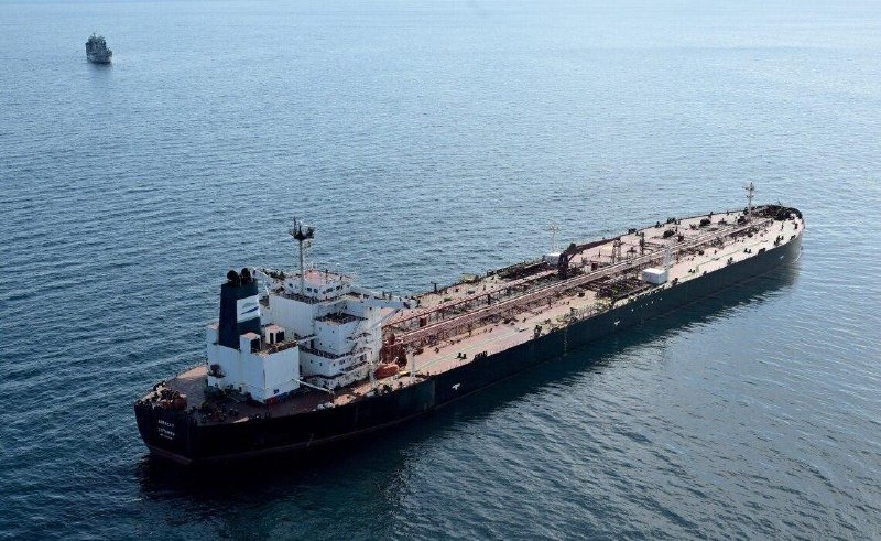 Aerial view of a large dark-hulled oil tanker with a white superstructure anchored in calm, open blue water, with a smaller vessel visible in the distance.