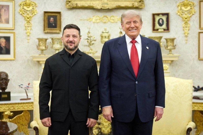 Two men pose smiling in an ornate room with gilded decor and framed portraits; one wears a dark suit, the other a navy suit with red tie.