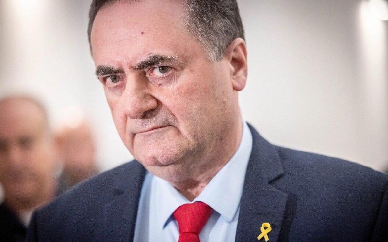 A man in a dark suit, light blue shirt, and red tie with a yellow ribbon pin stands indoors with a serious expression, looking off-camera.