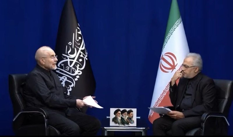 Two seated men face each other in a formal setting flanked by the Iranian flag and a black banner with white Arabic calligraphy, with a small table holding a framed portrait between them.