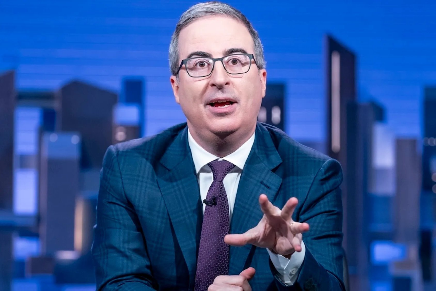 A gray-haired man in glasses, a blue plaid suit, and a purple tie gestures while speaking against a blue cityscape backdrop.