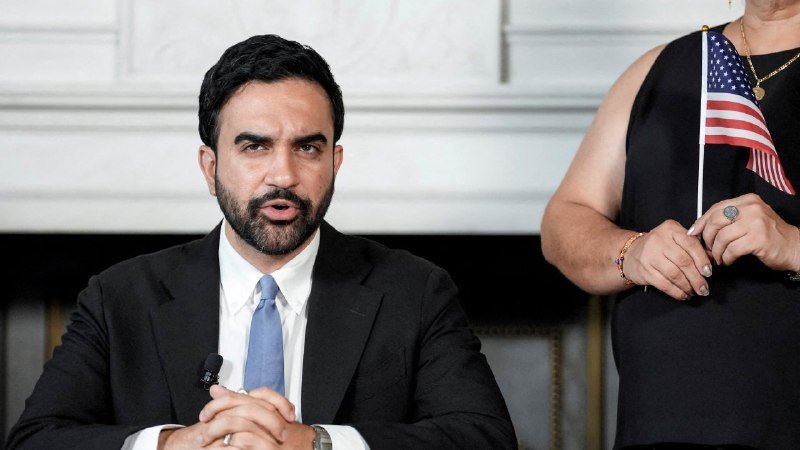 A bearded man in a black suit and blue tie speaks at a microphone indoors, while a woman beside him in a black dress holds a small American flag.