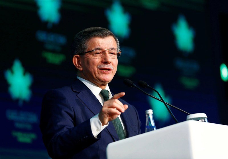 A bespectacled man in a dark suit and green tie speaks at a podium, gesturing with his hand against a backdrop of illuminated green leaf graphics.