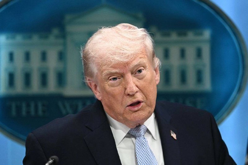 A blond man in a dark suit and light blue tie speaks at a podium, with a blue backdrop displaying the White House seal behind him.