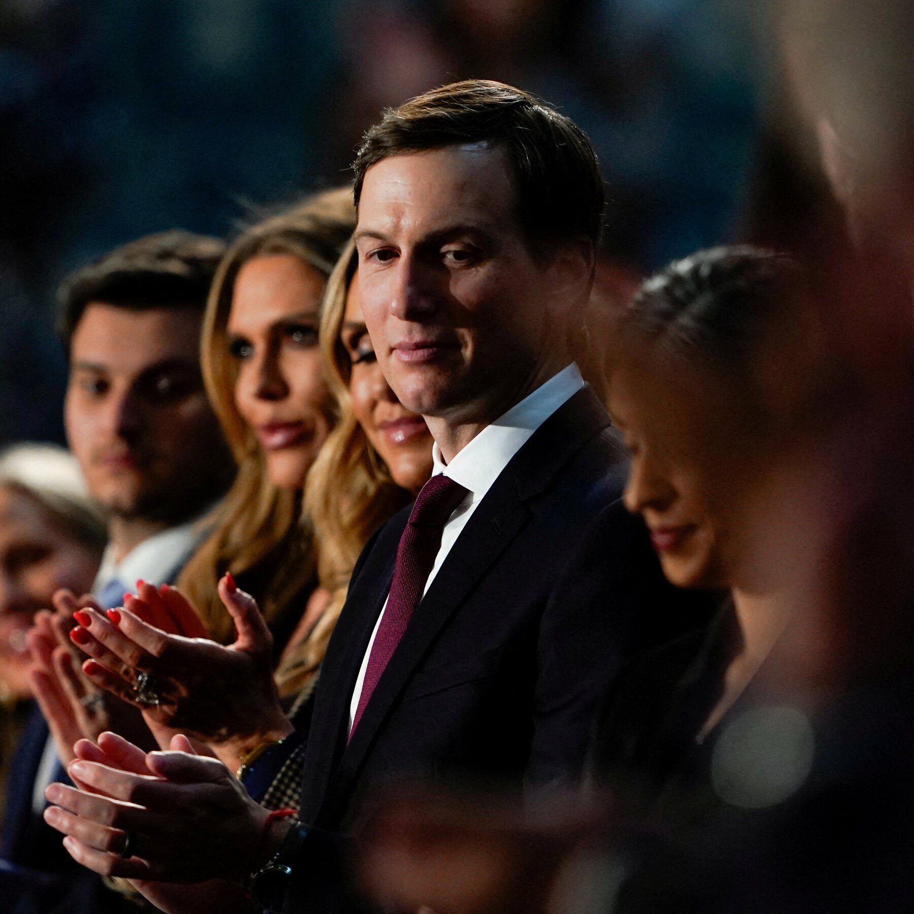 A man in a dark suit and burgundy tie stands among seated applauding people in a dimly lit indoor venue.