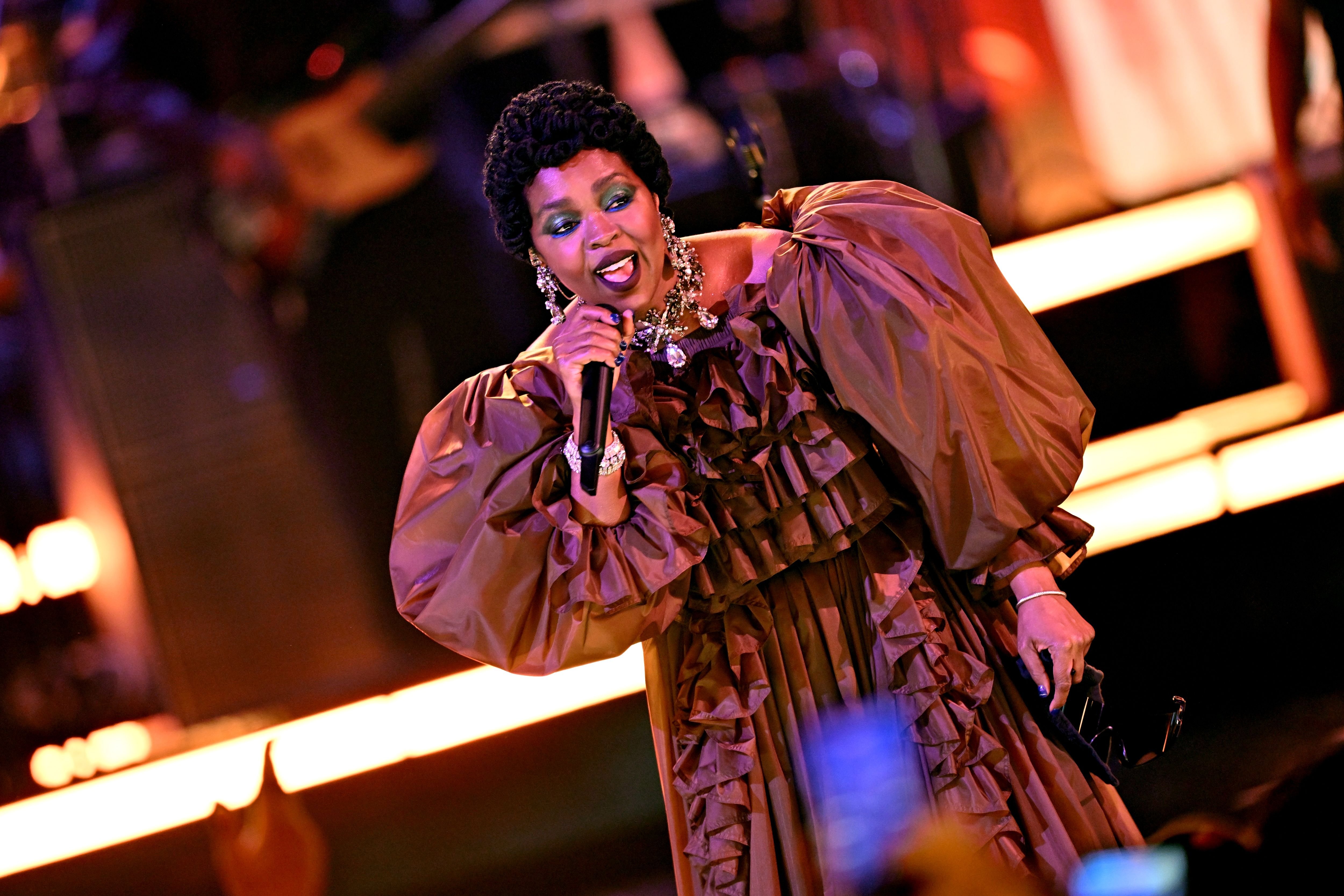 A woman in a ruffled bronze gown and statement jewelry sings into a microphone on a stage illuminated by warm orange lighting.