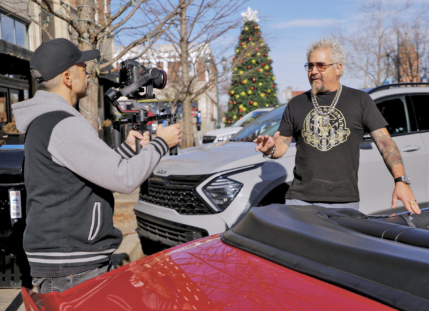 A cameraman films a man with spiky gray hair and tattoos beside parked cars on a street decorated with a lit Christmas tree.