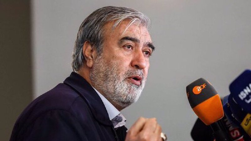 A political cartoon illustration shows a man with blonde hair in a blue suit wielding a hammer inside a coffin labeled "IRAN," with smoke surrounding the scene.