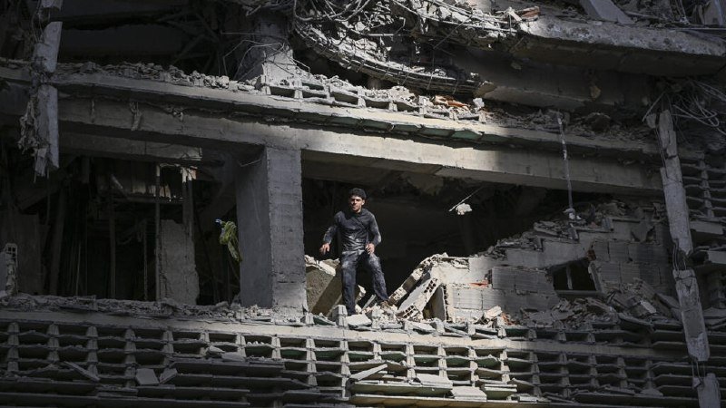 Numerous people climb and search through a large pile of concrete and brick rubble under a blue sky.